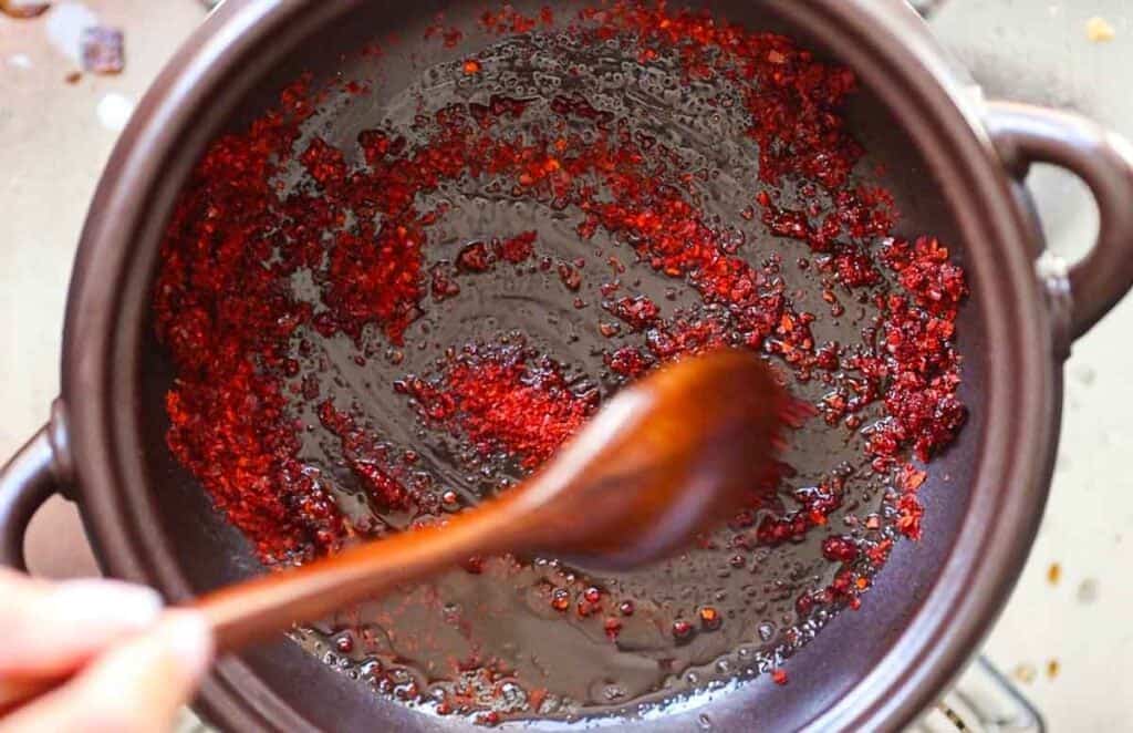 A hand is stirring gochugaru in a black pot with a wooden spoon. The paste is sizzling and spreading across the surface of the pot, creating a vibrant red swirl against the dark background.